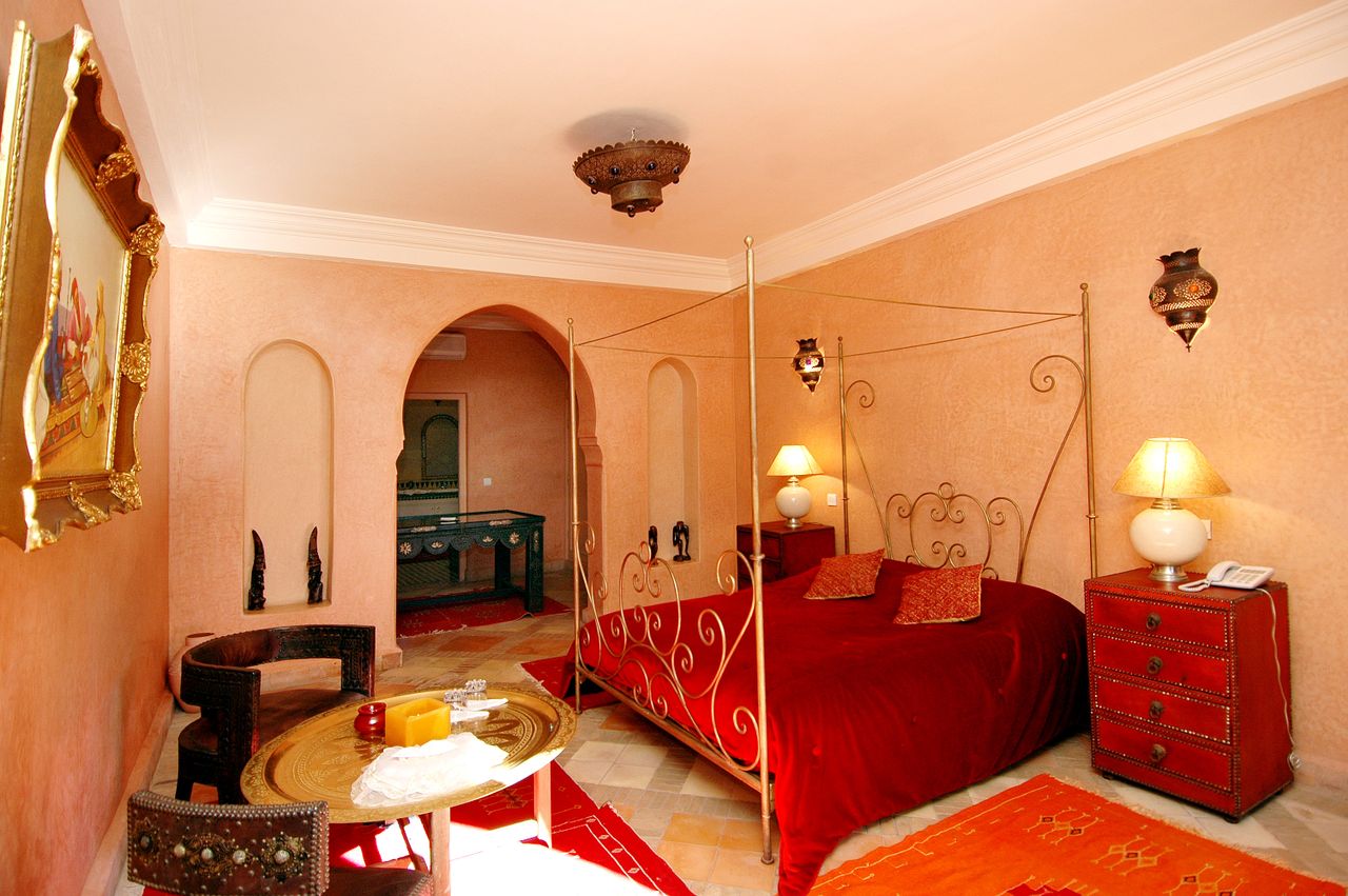 about-01 Luxurious Moroccan-themed bedroom featuring a red canopy bed, ornate decor, and warm lighting.
