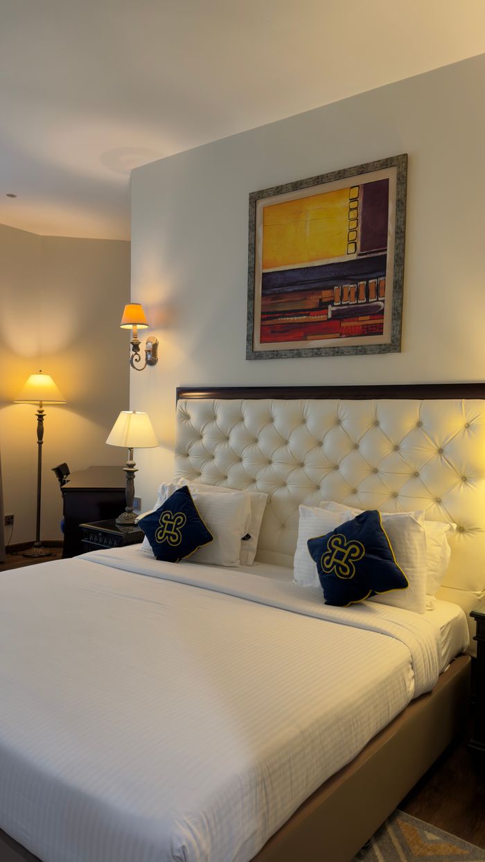 gallery-2 Luxurious hotel room featuring a plush bed, decorative pillows, and modern lighting for a cozy stay.